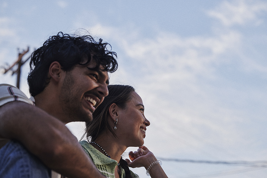 A man and a woman smiling and looking into the distance, standing close together outdoors under a blue sky with light clouds.