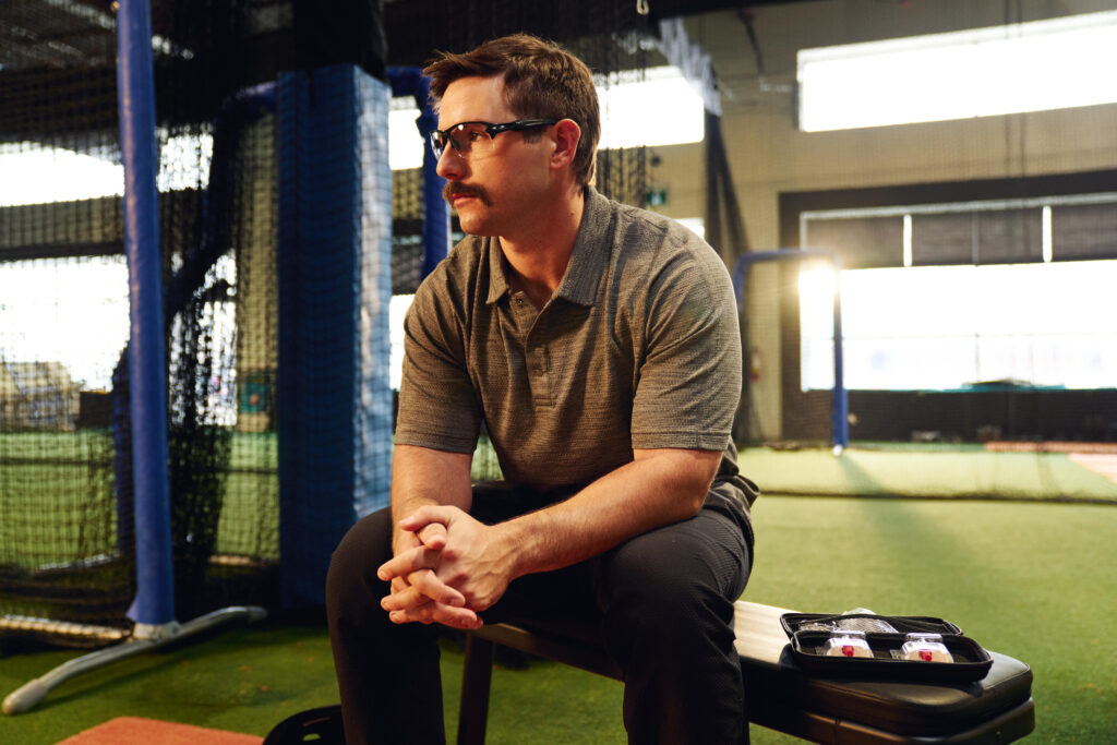 A man with glasses and a mustache sits on a bench in an indoor sports facility, hands clasped, wearing a gray polo shirt and black pants. A small open case with gear is on the bench beside him.