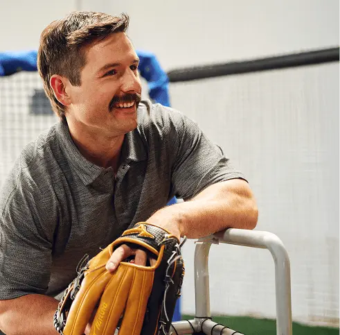 A man with a mustache wearing a gray polo shirt smiles while holding a baseball glove and leaning on a metal fence, with a netted area visible in the background.