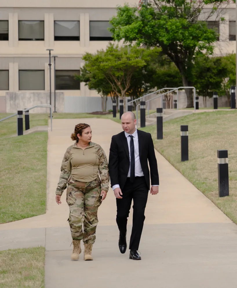 A woman in military uniform and a man in a black suit walk side by side on a paved path outside a building, with grass and trees in the background.