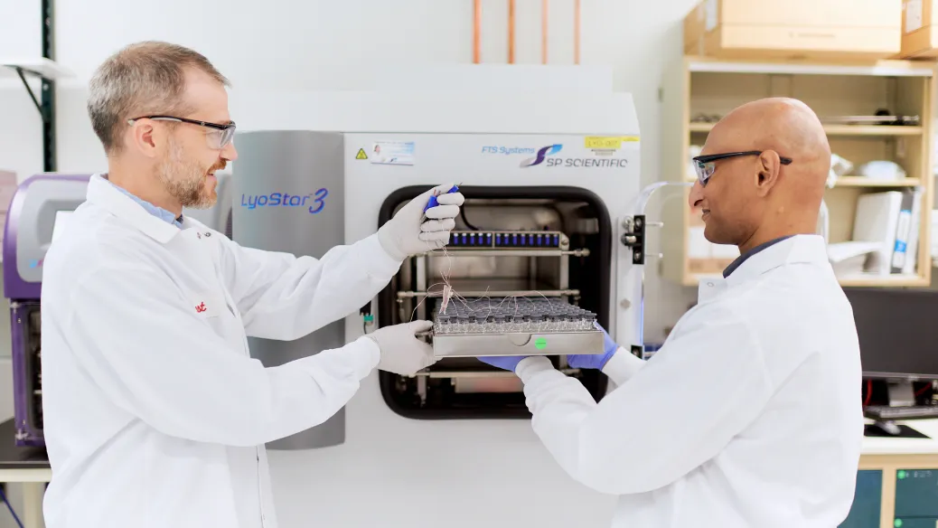 Two scientists in lab coats and gloves work together with laboratory equipment, one holding a tray of vials while the other uses a pipette, standing in front of a freeze dryer machine in a laboratory setting.