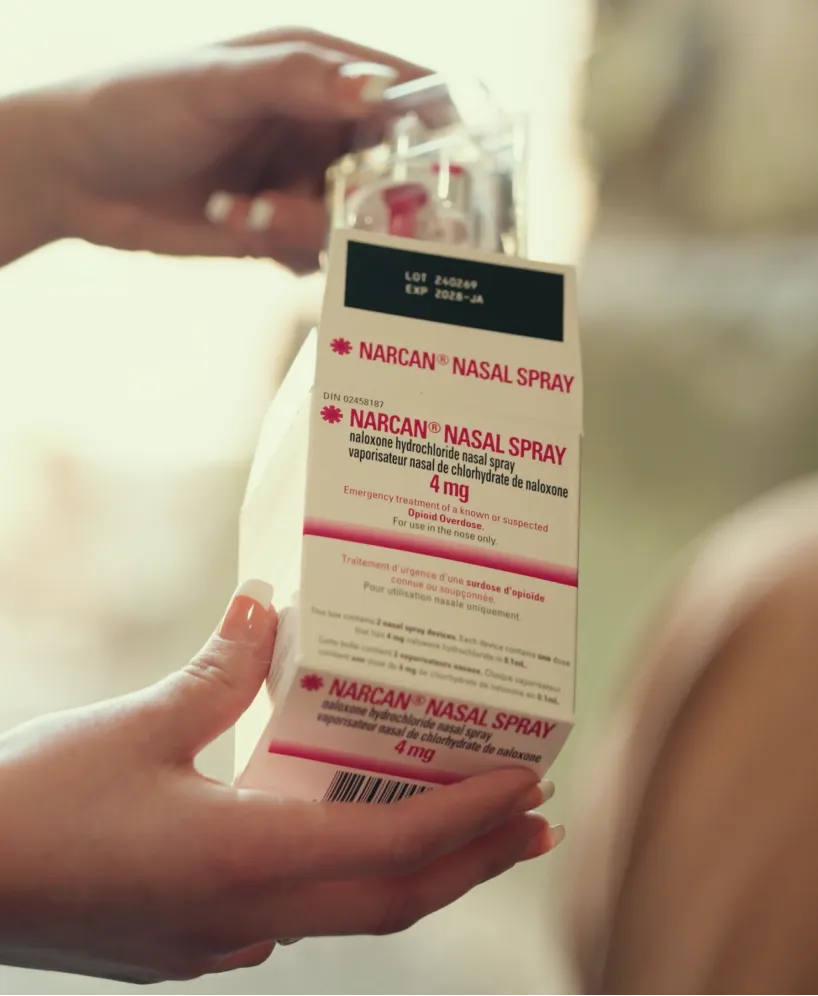A person holds a Narcan Nasal Spray box, used for emergency treatment of opioid overdoses. The packaging is white with red and black text, showing dosage and instructions.