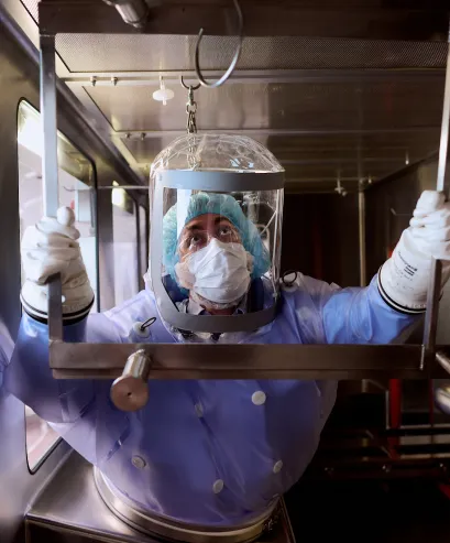 A person wearing a protective suit, gloves, face mask, and hair net works inside a laboratory, reaching up inside a containment area with a transparent shield surrounding their head.