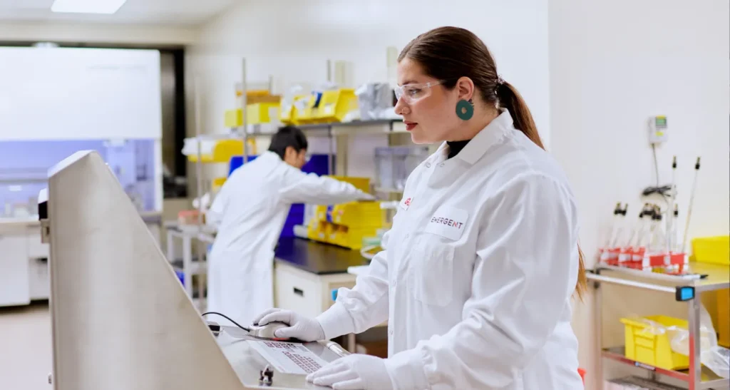 A scientist in a lab coat and safety glasses operates equipment in a laboratory, while another person works in the background near a bench with yellow storage bins and lab supplies.