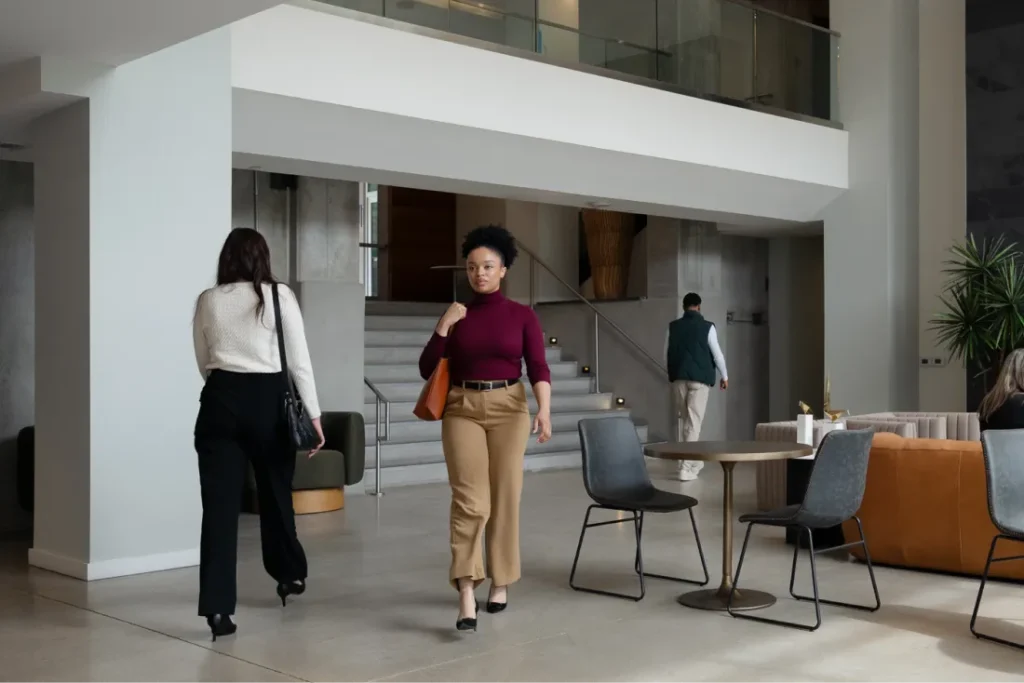 A woman in a maroon top and tan pants walks through a modern lobby. Another woman walks away, and a man ascends stairs. Modern furniture and plants decorate the space.