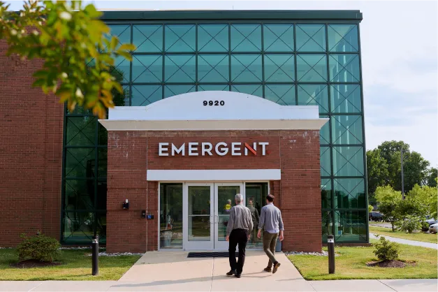 Two people walk toward the entrance of a brick building with glass windows. A sign above the door reads “EMERGENT,” and the number “9920” is above the entrance. Trees and bushes line the walkway.