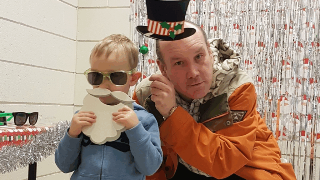 A child wearing sunglasses and holding a fake beard stands next to an adult in an orange coat, who holds a festive hat prop above his head. Both pose playfully in front of a decorated, tinsel-covered backdrop.