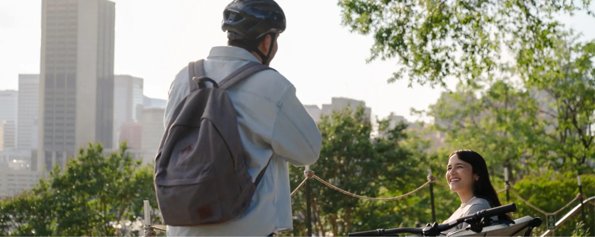 A person wearing a helmet and backpack stands with a bike, facing a smiling woman sitting on a bench outdoors in a park with city buildings in the background.