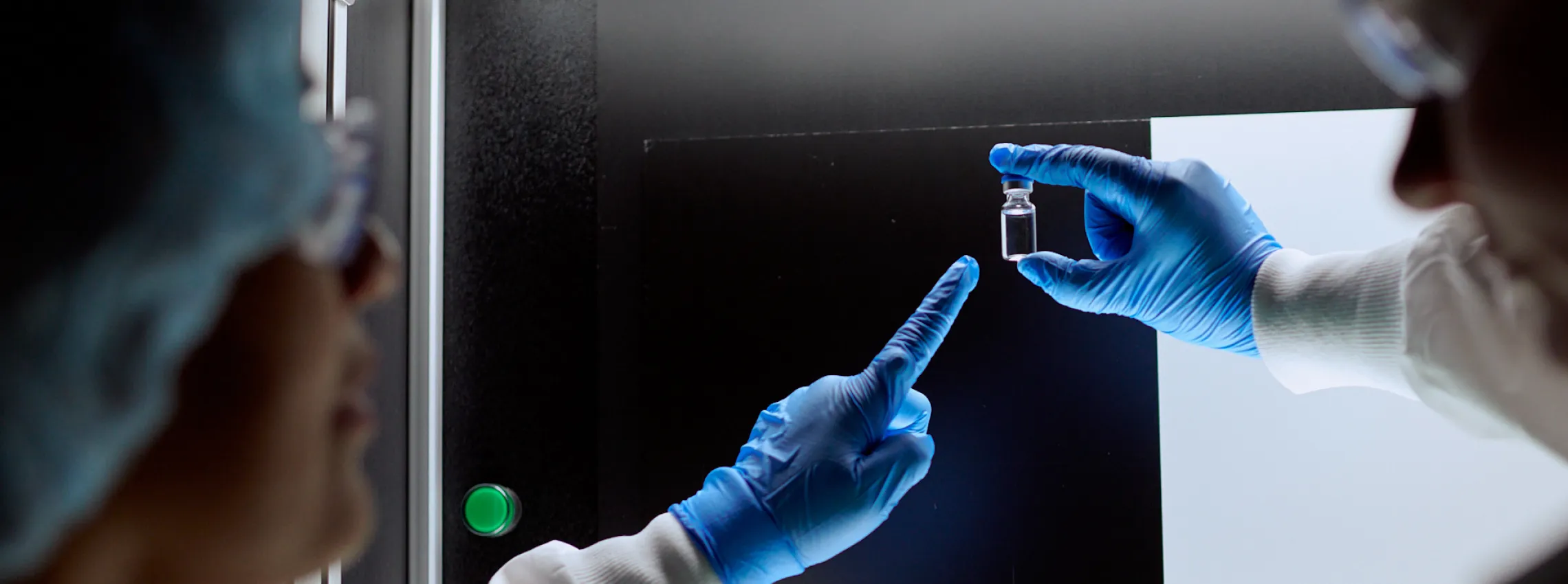 Two people wearing blue gloves and lab coats inspect a small clear vial against a dark background; one person points at the vial, indicating discussion or analysis.