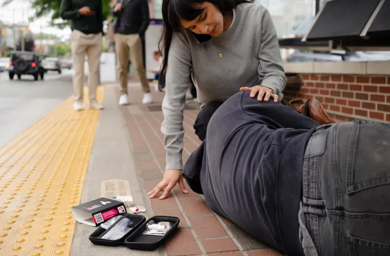 A person kneels by someone lying on a sidewalk, appearing to assist them. Open medical kits and naloxone boxes are visible nearby. Other people walk in the background.