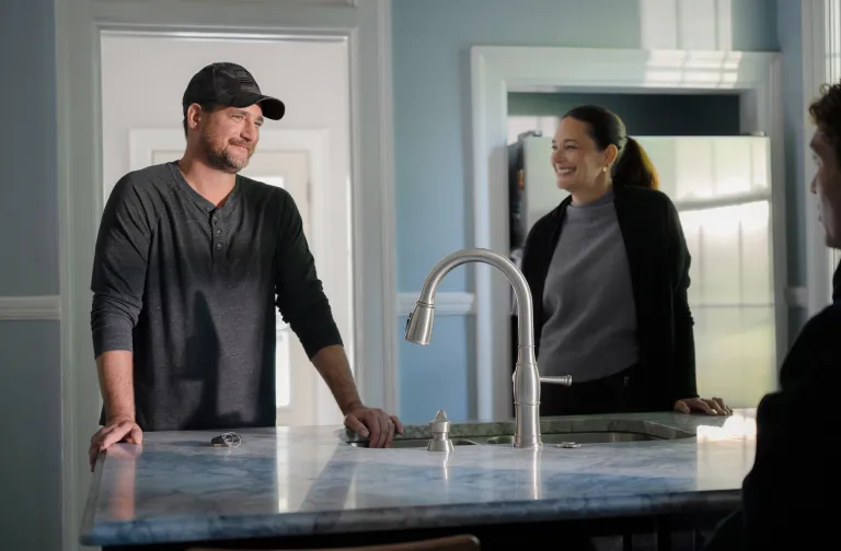 A man in a dark baseball cap and a woman in a gray sweater stand smiling by a marble kitchen island with a sink and faucet, talking to someone offscreen.
