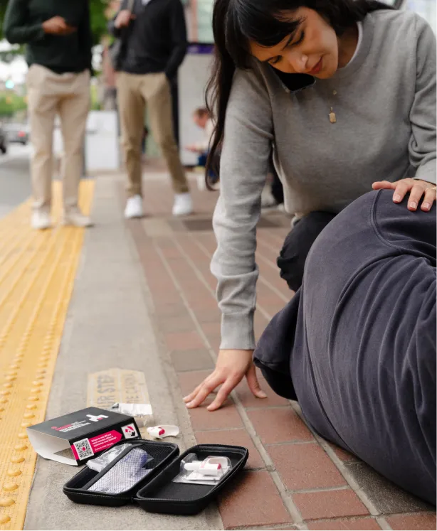 A woman kneels beside a person lying on a sidewalk. In the foreground, a naloxone kit is open, suggesting a medical emergency response. Other people stand blurred in the background.