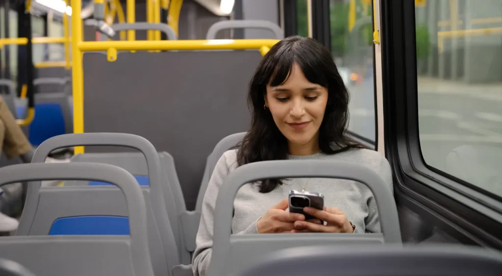 A woman with dark hair sits alone on a bus, smiling as she looks at her phone. The bus appears empty, and it’s daytime outside.
