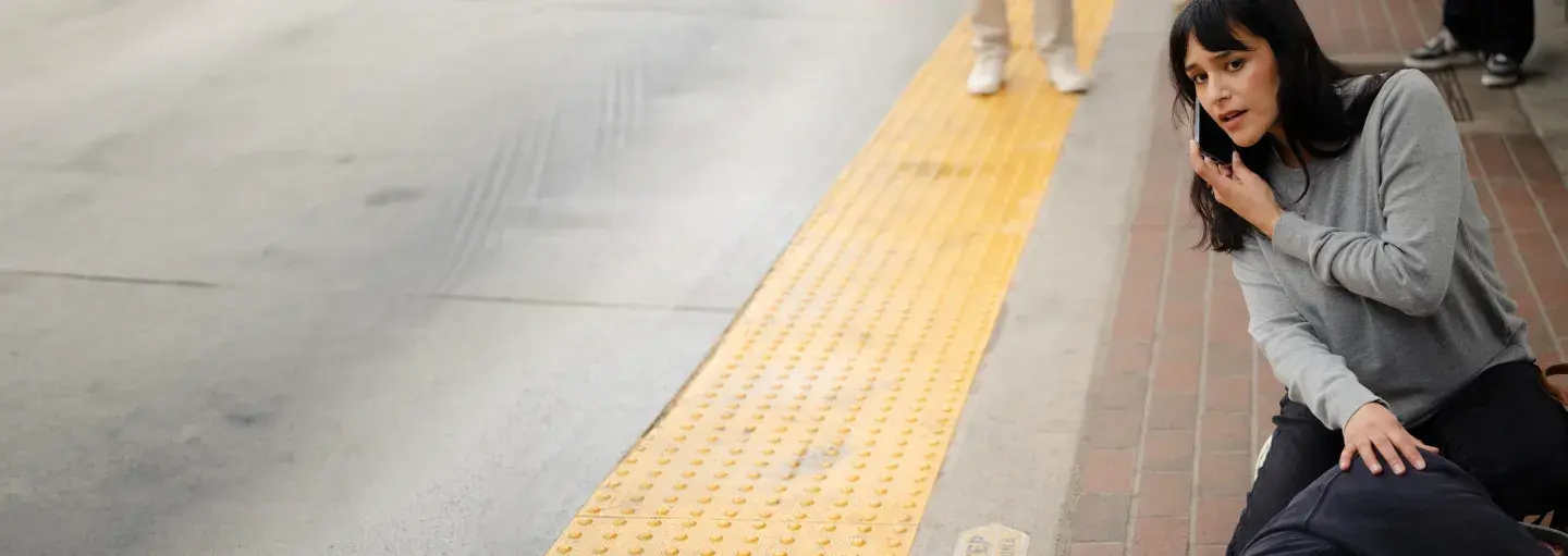 A woman kneels on a sidewalk next to a person lying down, checking their pulse and looking concerned. Yellow tactile paving marks the edge of the sidewalk near a street.